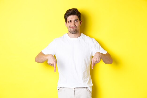 Młody nieszczęśliwy facet krzywiący się, wskazujący palcami w dół na reklamę, rozczarowany produktem, stojący na żółtym tle.