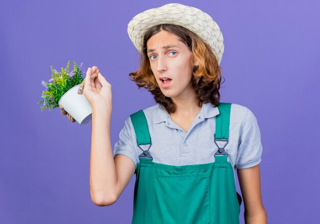 Młody ogrodnik człowiek ubrany w kombinezon i kapelusz gospodarstwa roślina doniczkowa jest zdezorientowany