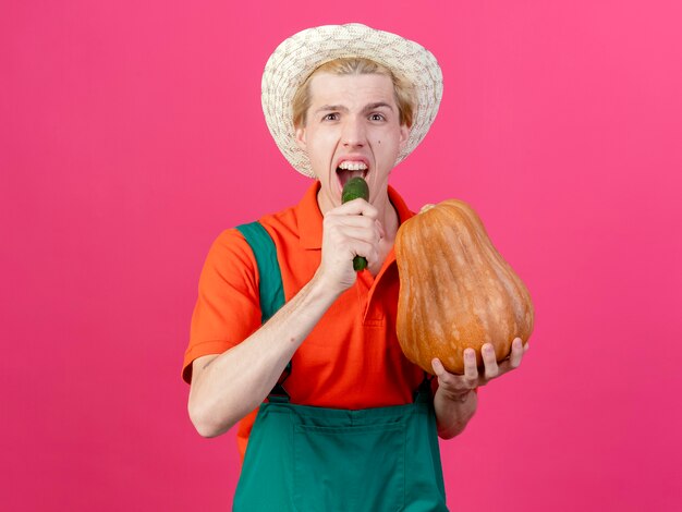 Młody ogrodnik mężczyzna na sobie kombinezon i kapelusz, trzymając dyni i ogórek