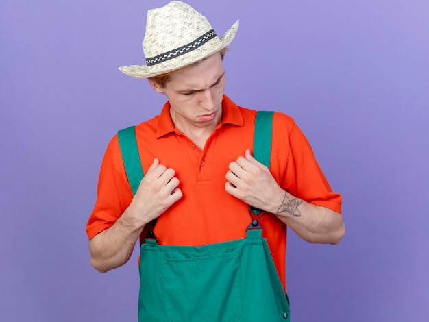 Bezpłatne zdjęcie młody ogrodnik mężczyzna ubrany w kombinezon i kapelusz, patrząc zdezorientowany