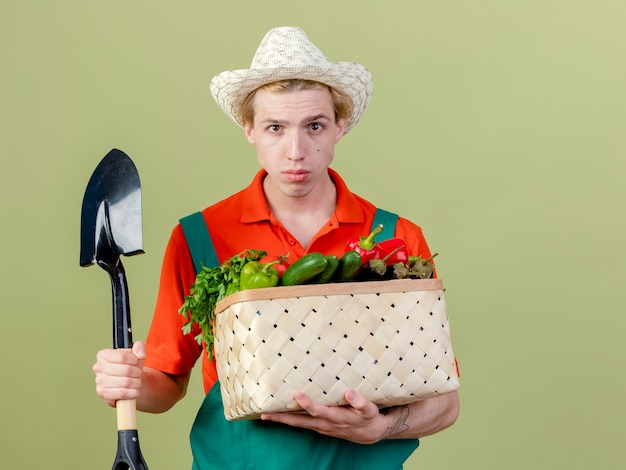 Młody ogrodnik mężczyzna ubrany w kombinezon i kapelusz trzymający skrzynię pełną warzyw i łopatę patrząc na kamerę z poważną twarzą stojącą na jasnym tle