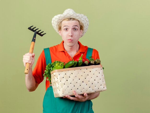 Młody ogrodnik mężczyzna ubrany w kombinezon i kapelusz trzymający skrzynię pełną warzyw i mini grabie patrząc na aparat zdezorientowany stojąc na jasnym tle
