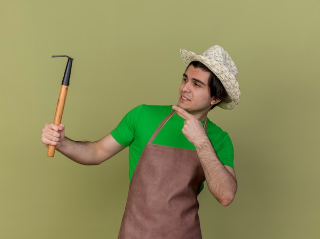 Bezpłatne zdjęcie młody ogrodnik mężczyzna w fartuchu i kapeluszu, trzymając mini grabie, wskazując palcem wskazującym na niego uśmiechnięty wesoło stojąc nad jasną ścianą