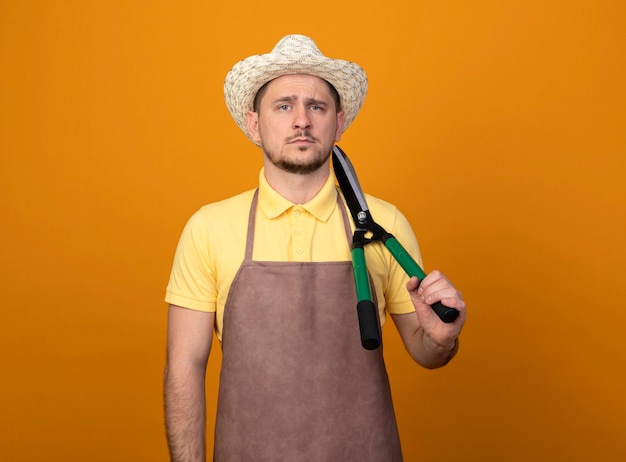 Bezpłatne zdjęcie młody ogrodnik mężczyzna w kombinezon i kapelusz, trzymając nożyce do żywopłotu na ramieniu z poważnym wyrazem pewności