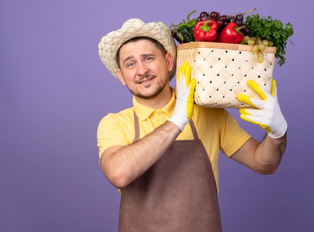 Młody ogrodnik w kombinezonie i kapeluszu w roboczych rękawiczkach trzymający skrzynię pełną warzyw na ramieniu, patrząc z przodu z uśmiechem na twarzy stojącej nad fioletową ścianą