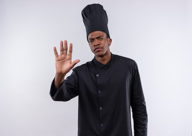 Młody pewny siebie afro-amerykański kucharz w gestach munduru szefa kuchni stop znak ręką na białym tle na białym tle z miejsca na kopię