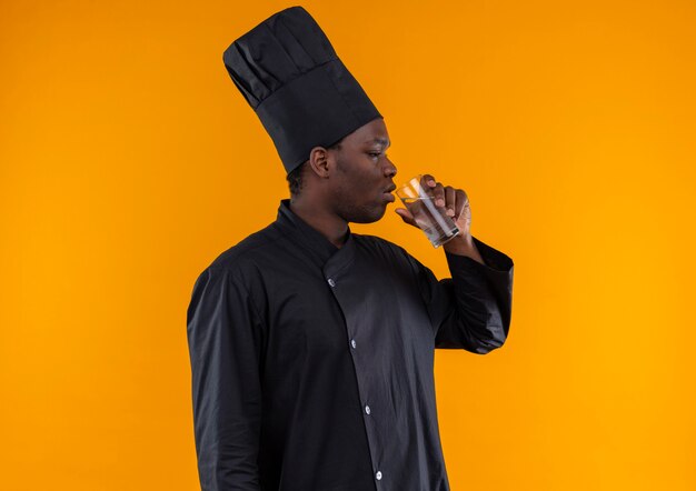 Młody pewny siebie afro-amerykański kucharz w mundurze szefa kuchni stoi bokiem i pije szklankę wody na białym tle na pomarańczowym tle z miejsca na kopię