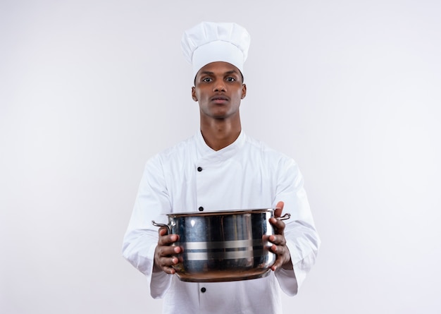 Młody pewny siebie afro-amerykański kucharz w mundurze szefa kuchni trzyma rondel obiema rękami na na białym tle z miejsca na kopię