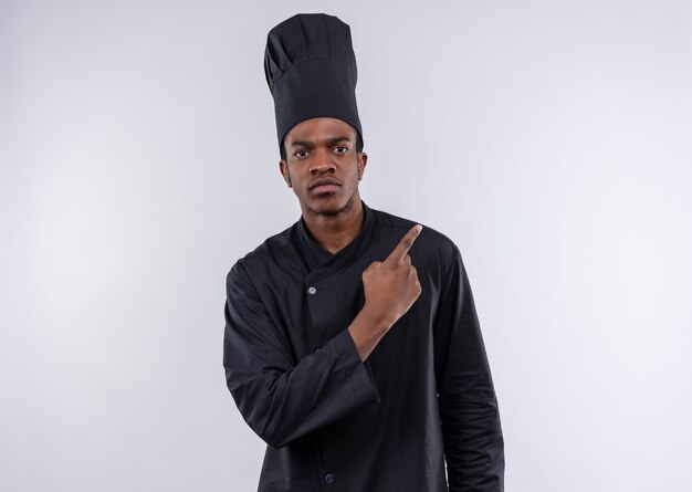 Młody pewny siebie afro-amerykański kucharz w mundurze szefa kuchni wskazuje na bok na białym tle na białym tle z miejsca na kopię