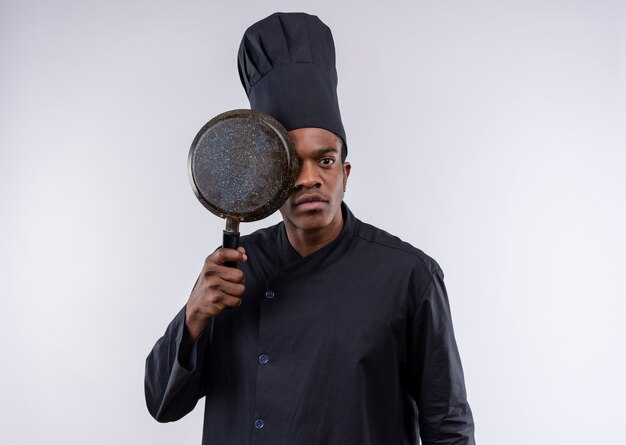 Młody pewny siebie afro-amerykański kucharz w mundurze szefa kuchni zamyka oko z patelni na białym tle na białym tle z miejsca na kopię