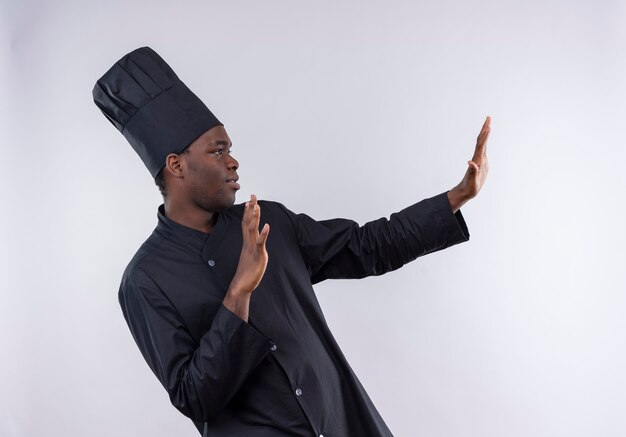 Młody pewny siebie afroamerykański kucharz w mundurze szefa kuchni udaje, że broni rękami na białym tle z miejsca na kopię
