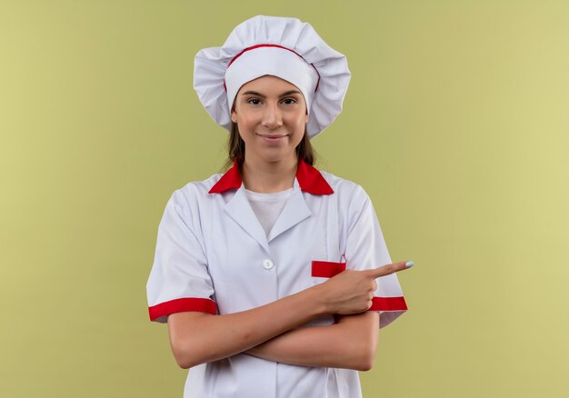 Młody pewny siebie kucharz kaukaski dziewczyna w mundurze szefa kuchni krzyżuje ramiona i wskazuje na bok na zielono z miejsca na kopię