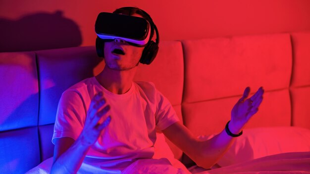 Młody pod wrażeniem mężczyzna w okularach wirtualnej rzeczywistości z niebiesko-czerwonym podświetleniem w pokoju na łóżku. Rozrywka w domu