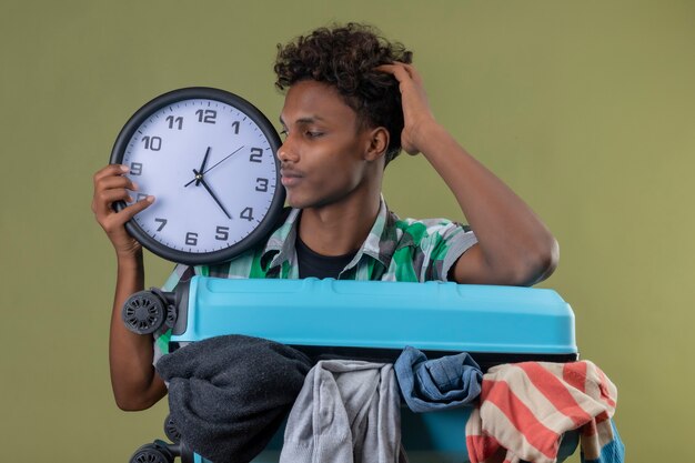 Młody podróżnik african american człowiek stojący z walizką pełną ubrań trzyma zegar patrząc na niego z zmieszanym wyrazem twarzy na zielonym tle