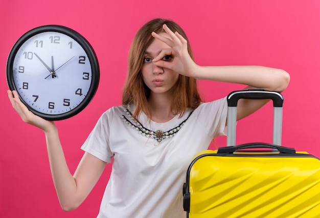 Bezpłatne zdjęcie młody podróżnik dziewczyna trzyma zegar robi gest patrzenia i kładzie rękę na walizce na odizolowanej różowej ścianie