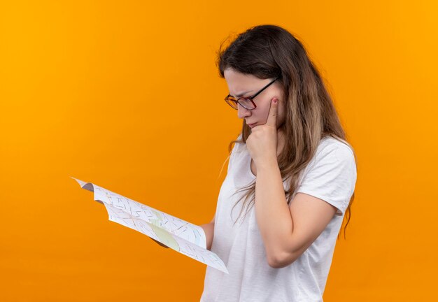 Młody podróżnik kobieta w białej koszulce trzymając mapę patrząc na to z zamyślonym wyrazem twarzy myślenia stojącego nad pomarańczową ścianą