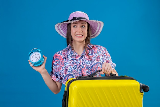Bezpłatne zdjęcie młody podróżnik kobieta w kapeluszu lato stojący z żółtą walizką trzymając budzik zestresowany i nerwowy na niebieskim tle
