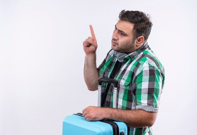 Młody podróżnik mężczyzna w kraciastej koszuli, trzymając walizkę wskazując palcem w bok, patrząc zdezorientowany stojąc na białej ścianie