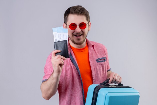 Młody podróżnik mężczyzna w okularach przeciwsłonecznych, trzymając bilety lotnicze i walizkę, patrząc na kamery szczęśliwy i pozytywny, uśmiechnięty troskliwie stojący na białym tle