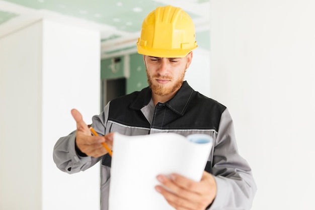 Bezpłatne zdjęcie młody poważny inżynier w stroju roboczym i żółtym kasku, w zamyśleniu patrzący na plan nowego mieszkania w pracy
