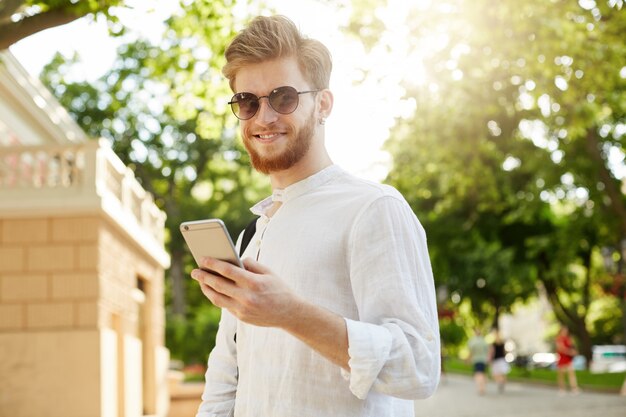 Młody pozytywny i uśmiechnięty rudowłosy mężczyzna z brodą i kolczykiem w okularach przeciwsłonecznych, patrząc przez sieci społecznościowe na swoim smartfonie w drodze do domu.