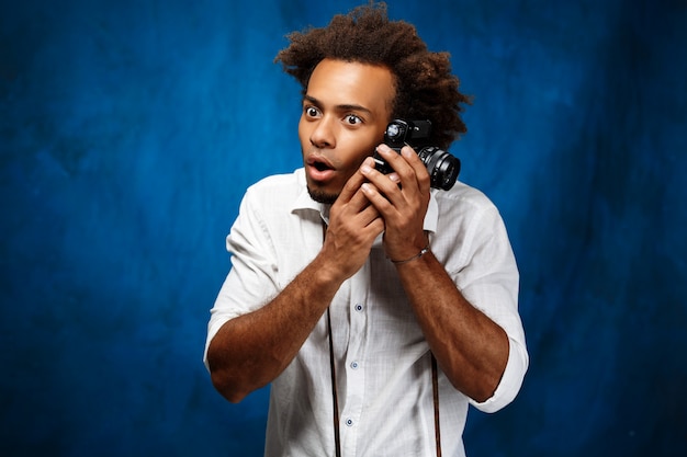 Młody przystojny afrykański mężczyzna trzyma starą kamerę nad błękit ścianą.