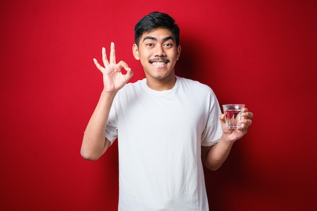 Młody Przystojny Azjatycki Mężczyzna Ubrany W Białą Koszulkę Pije Szklankę Wody, Wskazując Kciukiem W Górę Do Boku, Uśmiechając Się Zadowolony Z Otwartymi Ustami Na Czerwonym Tle Premium Zdjęcia