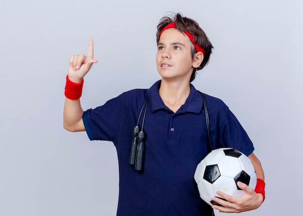 Młody przystojny chłopak sportowy w opasce i opaskach na nadgarstek z szelkami dentystycznymi i skakanką wokół szyi, trzymając piłkę nożną patrząc w górę, wykonując gest przegranego na białym tle