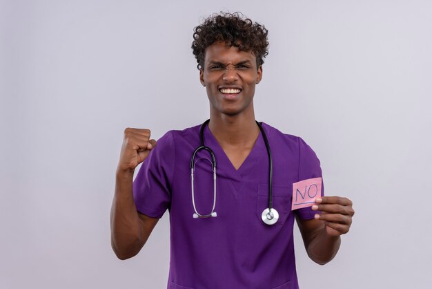 Młody przystojny ciemnoskóry lekarz z kręconymi włosami w fioletowym mundurze ze stetoskopem pokazujący papierową kartę z napisem nie z zaciśniętą pięścią