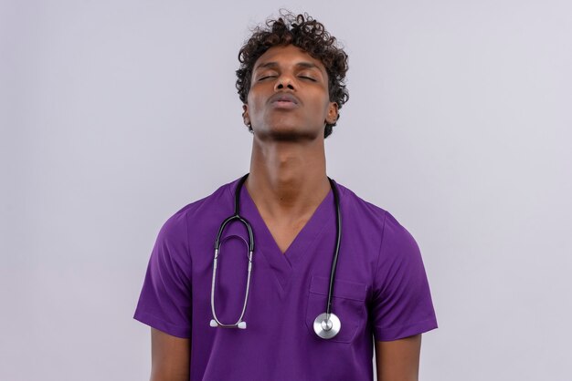 Młody przystojny, ciemnoskóry lekarz z kręconymi włosami w fioletowym mundurze ze stetoskopem zamykającym oczy ze zmęczonym wyrazem