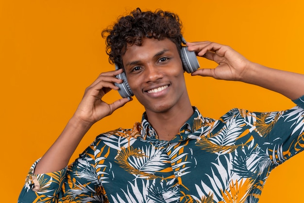Młody przystojny ciemnoskóry mężczyzna z kręconymi włosami w liściach drukowaną koszulę w słuchawkach uśmiechnięty, trzymając ręce na słuchawkach