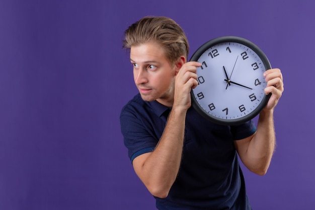 Młody przystojny facet trzyma zegar patrząc na bok z pewnym siebie wyrazem stojącym na fioletowym tle