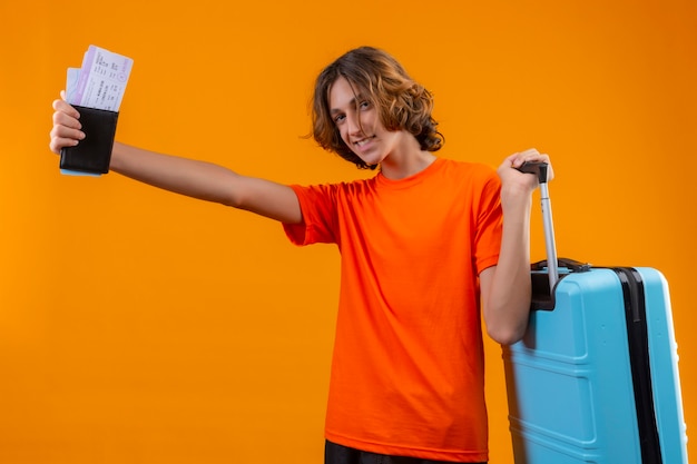 Młody przystojny facet w pomarańczowej koszulce stojącej z walizką podróżną, trzymając bilety lotnicze, uśmiechając się wesoło pozytywnie i szczęśliwie na żółtym tle
