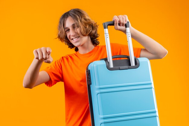 Młody przystojny facet w pomarańczowej koszulce trzyma walizkę podróżną, wskazując palcem na aparat, uśmiechając się wesoło wyglądając szczęśliwie i pozytywnie stojąc na żółtym tle