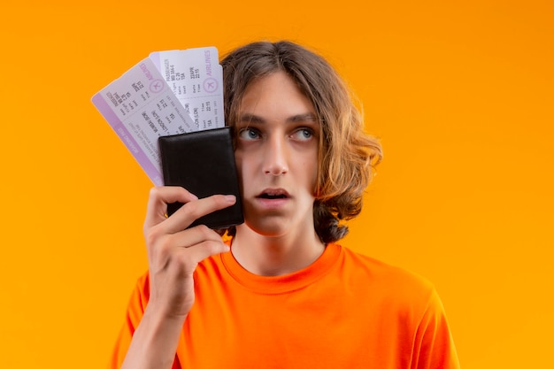 Młody przystojny facet w pomarańczowej koszulce trzymając bilety lotnicze patrząc na bok zdezorientowany ze smutnym wyrazem twarzy