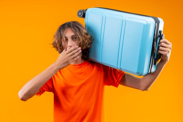 Młody przystojny facet w pomarańczowej koszulce, trzymając walizkę podróżną, patrząc zaskoczony i zdumiony, zakrywając usta ręką na żółtym tle