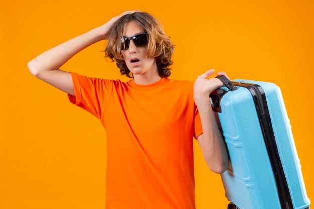 Młody przystojny facet w pomarańczowej koszulce w czarnych okularach przeciwsłonecznych, trzymając walizkę podróżną, stojąc z ręką na głowie za błąd wyglądający na zdezorientowanego pamiętaj o błędzie na żółtym tle