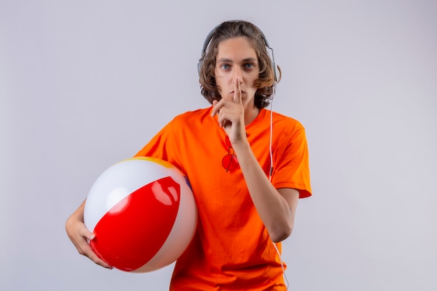 Bezpłatne zdjęcie młody przystojny facet w pomarańczowej koszulce ze słuchawkami, trzymając nadmuchiwaną piłkę, czyniąc gest ciszy z palcem na ustach stojących na białym tle