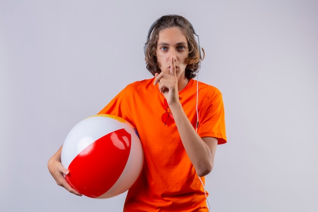 Młody przystojny facet w pomarańczowej koszulce ze słuchawkami, trzymając nadmuchiwaną piłkę robi gest ciszy z palcem na ustach stojących