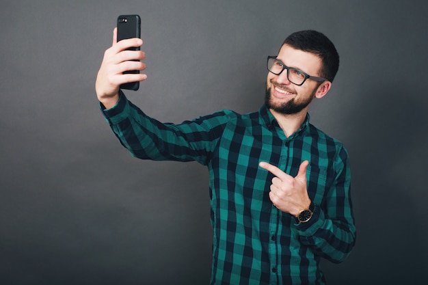 Młody przystojny hipster brodaty mężczyzna trzyma smartfon w ręce, zielona kraciasta koszula, pozytywne emocje, szczęśliwy, uśmiechnięty, szare tło