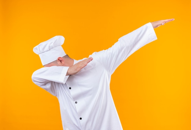 Bezpłatne zdjęcie młody przystojny kucharz w mundurze szefa kuchni kładzie głowę na ramieniu i podnosi inne ramię na białym tle na pomarańczowej przestrzeni