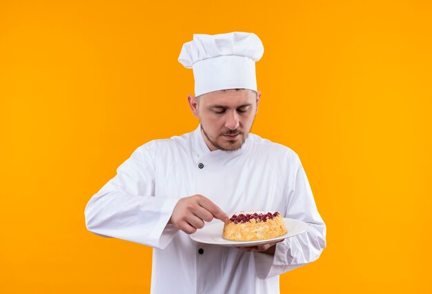 Młody Przystojny Kucharz W Mundurze Szefa Kuchni, Trzymając I Patrząc Na Talerz Ciasta Na Białym Tle Na Pomarańczowej Przestrzeni