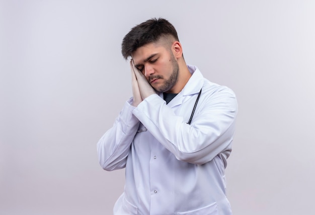Młody przystojny lekarz ubrany w białą suknię medyczną białe rękawiczki medyczne i stetoskop chce spać stojąc nad białą ścianą