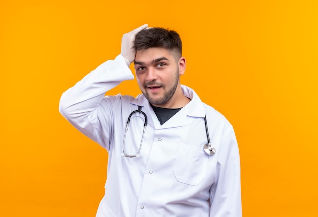 Bezpłatne zdjęcie młody przystojny lekarz ubrany w białą suknię medyczną białe rękawiczki medyczne i stetoskop głowę z ręką patrzącą z zapomnianą twarzą stojącą nad pomarańczową ścianą
