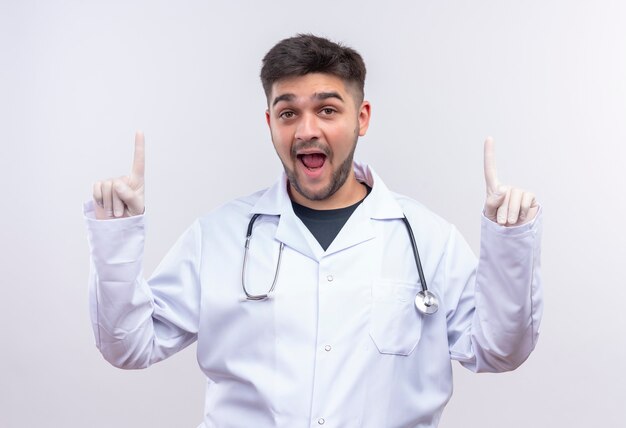 Młody przystojny lekarz ubrany w białą suknię medyczną białe rękawiczki medyczne i stetoskop patrząc w górę z palcami wskazującymi stojącymi nad białą ścianą