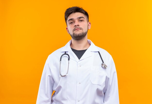 Młody Przystojny Lekarz Ubrany W Białą Suknię Medyczną Białe Rękawiczki Medyczne I Stetoskop Poważnie Patrząc Stojąc Na Pomarańczowej ścianie