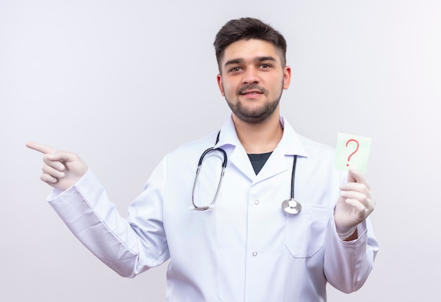 Młody przystojny lekarz ubrany w białą suknię medyczną białe rękawiczki medyczne i stetoskop uśmiechnięty, trzymając znak zapytania i wskazujący na prawo stojącego nad białą ścianą