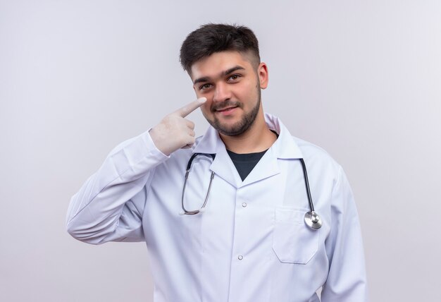 Młody przystojny lekarz ubrany w białą suknię medyczną białe rękawiczki medyczne i stetoskop uśmiechnięty, wskazując na nos stojąc na białej ścianie