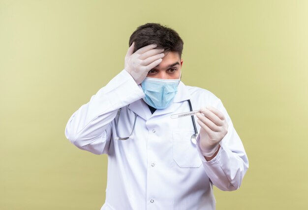 Młody przystojny lekarz ubrany w niebieską maskę medyczną białą suknię medyczną białe rękawiczki medyczne i stetoskop przestraszony trzymając termometr stojący nad ścianą khaki