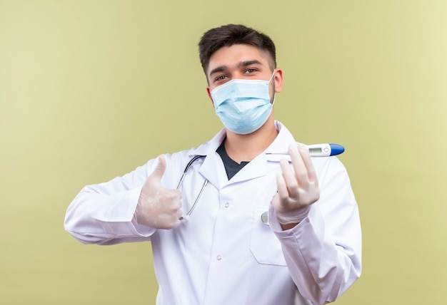 Młody przystojny lekarz ubrany w niebieską maskę medyczną białą suknię medyczną białe rękawiczki medyczne i stetoskop trzymający elektroniczny termometr robi radosne kciuki stojąc nad backgrou khaki